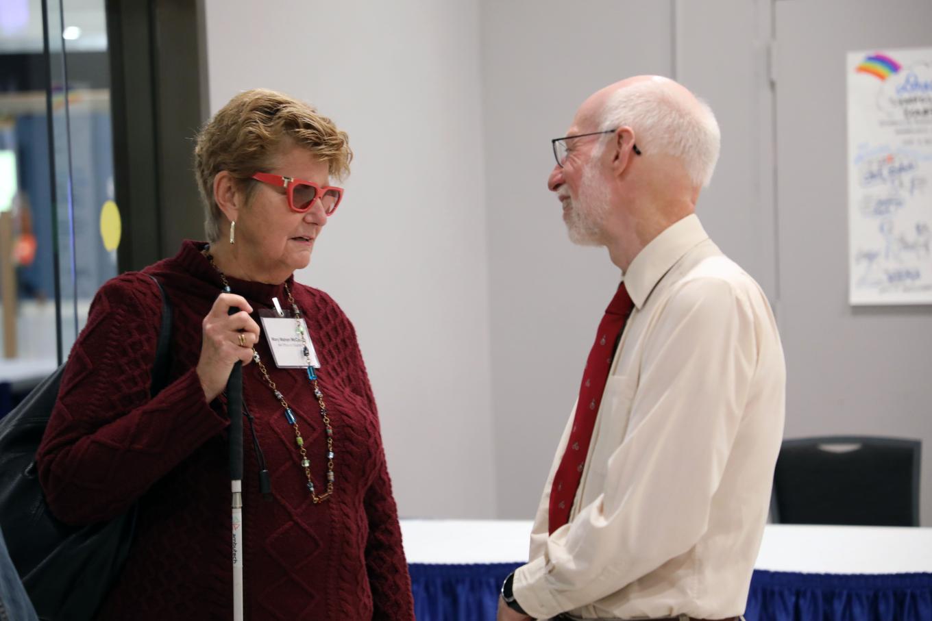 Mary Mahon McCauley, executive director of the Massachusetts Office on Disability, who is holding a white cane and wearing eyeglasses with a red frame and a red sweater, speaks to Bill Erickson from the Yang-Tan Institute. Bill is listening and wearing a red tie over a white business shirt.