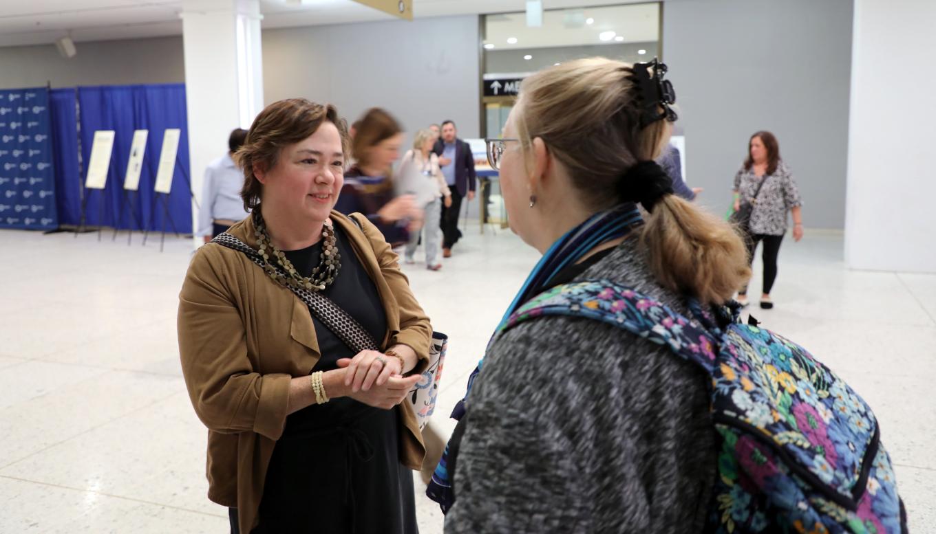 Yang-Tan Institute executive director Wendy Strobel Gower smiles while chatting with a DREAM attendee. Strobel Gower is professionally dressed in chunky jewelry, black dress and tan wrap; the attendee is wearing a blue scarf over a gray top and a backpack with a floral design.