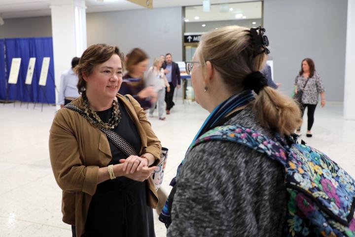 Yang-Tan Institute executive director Wendy Strobel Gower smiles while chatting with a DREAM attendee. Strobel Gower is professionally dressed in chunky jewelry, black dress and tan wrap; the attendee is wearing a blue scarf over a gray top and a backpack with a floral design.