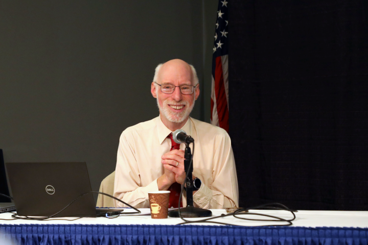 Bill Erickson seated at a speaker’s table while giving a presentation. He is wearing a white button-up dress shirt with a red tie. He is seated in front of an American flag and behind a laptop, mic, and coffee cup.